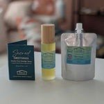 Three items from the brand 'Sacred Sweetgrass' are displayed on a white surface: a dark blue card, a glass bottle with a bamboo cap containing yellow spray and a silver resealable pouch with a white cap.