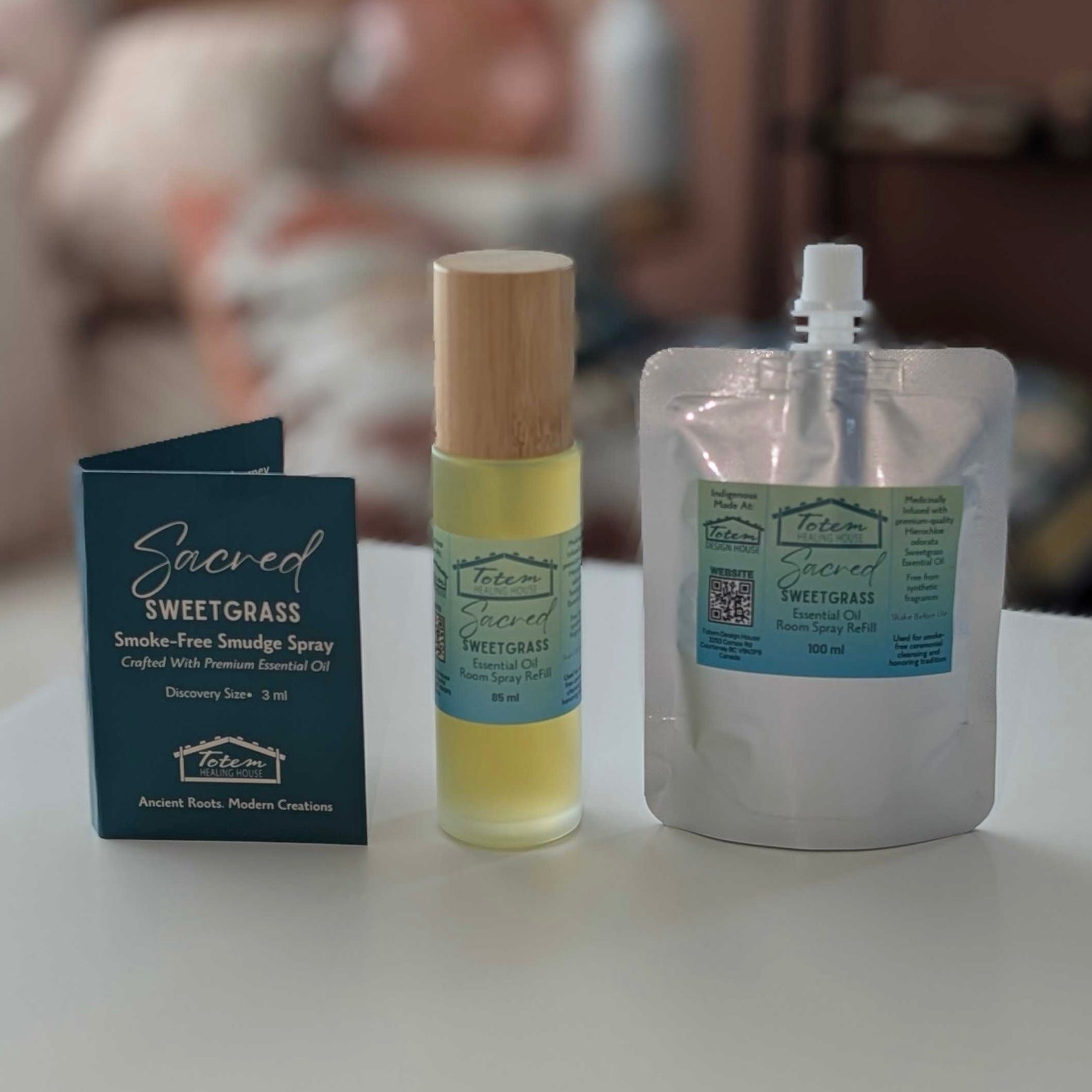 Three items from the brand 'Sacred Sweetgrass' are displayed on a white surface: a dark blue card, a glass bottle with a bamboo cap containing yellow spray and a silver resealable pouch with a white cap.